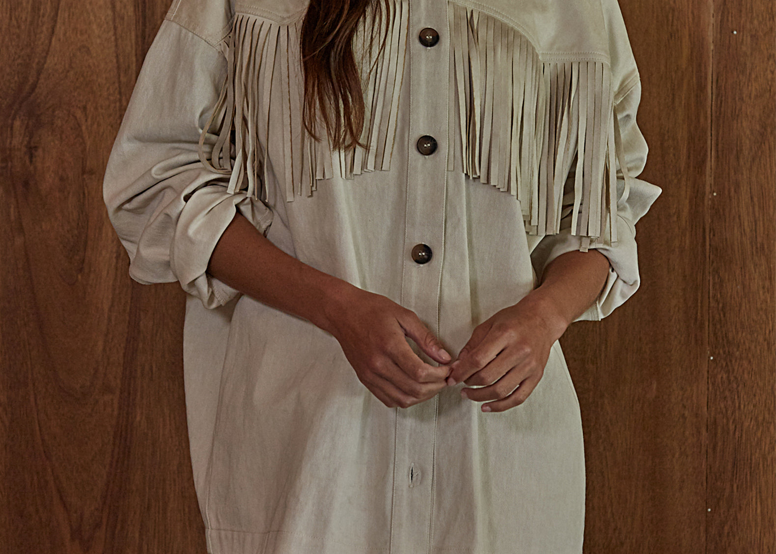 A woman wearing a light beige fringed shirt with a button-down front, standing against a dark wood-paneled wall.