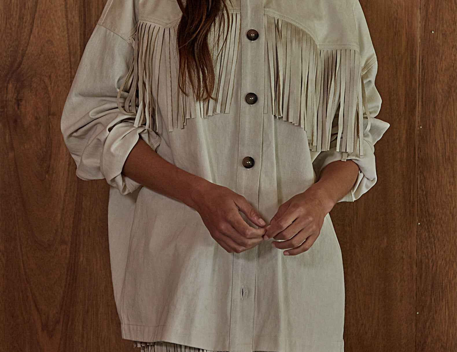 A woman wearing a light beige fringed shirt with a button-down front, standing against a dark wood-paneled wall.