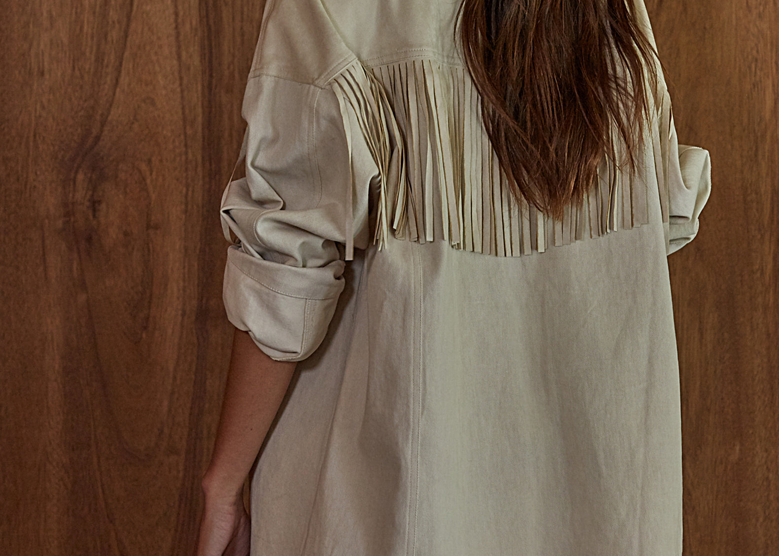 A woman shown from the back wearing an oversized light beige fringed jacket against a warm wood plank background.