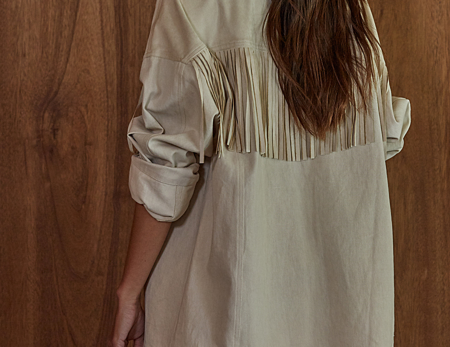 A woman shown from the back wearing an oversized light beige fringed jacket against a warm wood plank background.