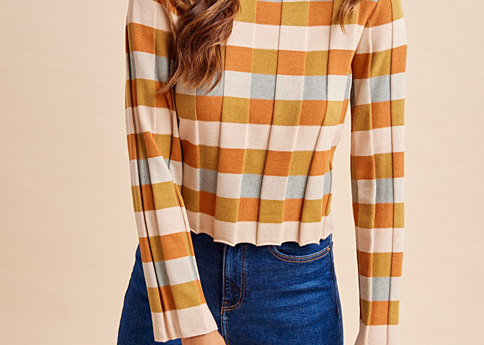 A woman wearing a long-sleeve cropped knit top with a warm-toned check pattern in orange, mustard, cream, and light blue, paired with dark blue jeans, against a solid beige background.