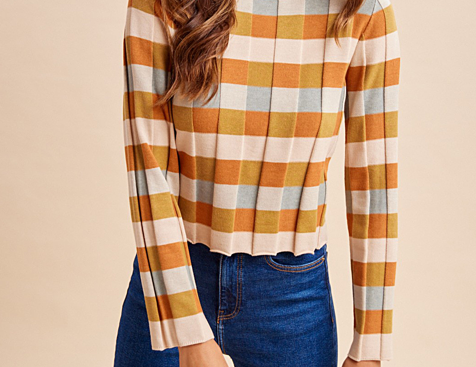 A woman wearing a long-sleeve cropped knit top with a warm-toned check pattern in orange, mustard, cream, and light blue, paired with dark blue jeans, against a solid beige background.