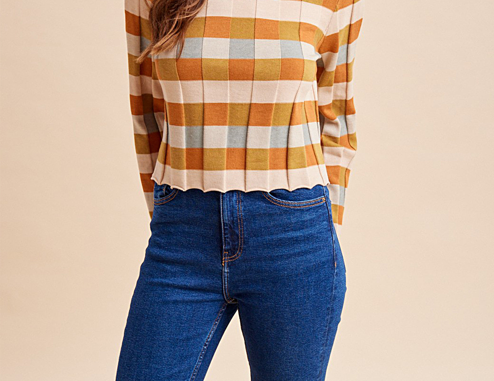 A woman wearing a long-sleeve knit top with a mustard yellow, orange, and cream plaid pattern, paired with blue jeans, against a solid beige background.