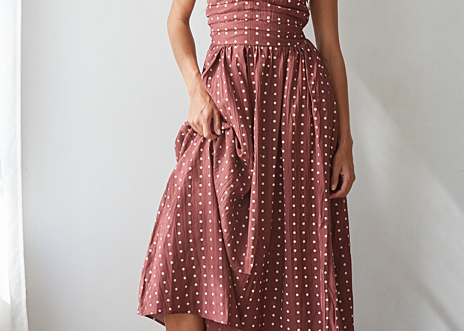 A woman models a strapless dusty rose pink midi dress with white polka dots, featuring a smocked bodice and an asymmetrical draped hem, paired with brown block-heel sandals against a white wall.