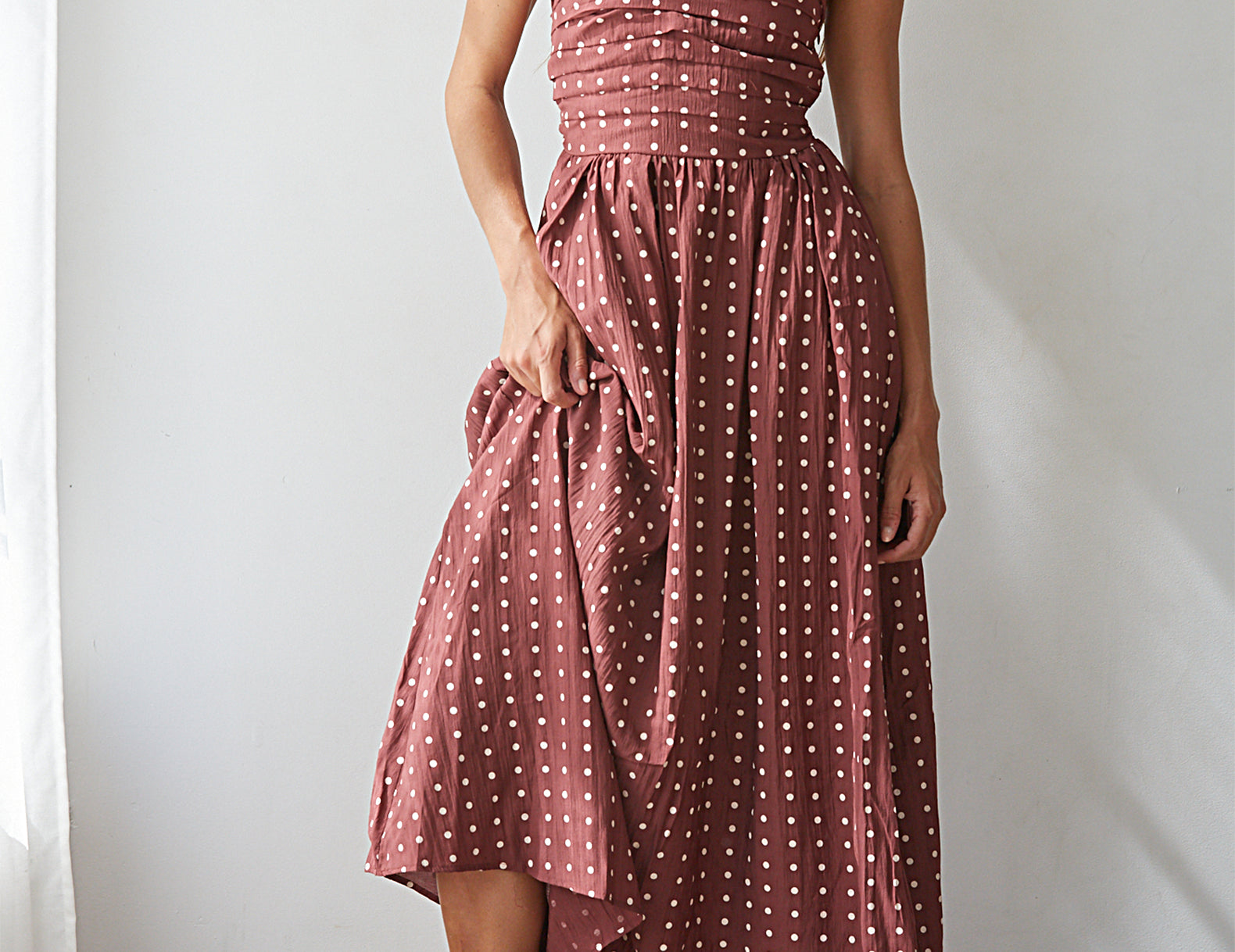 A woman models a strapless dusty rose pink midi dress with white polka dots, featuring a smocked bodice and an asymmetrical draped hem, paired with brown block-heel sandals against a white wall.