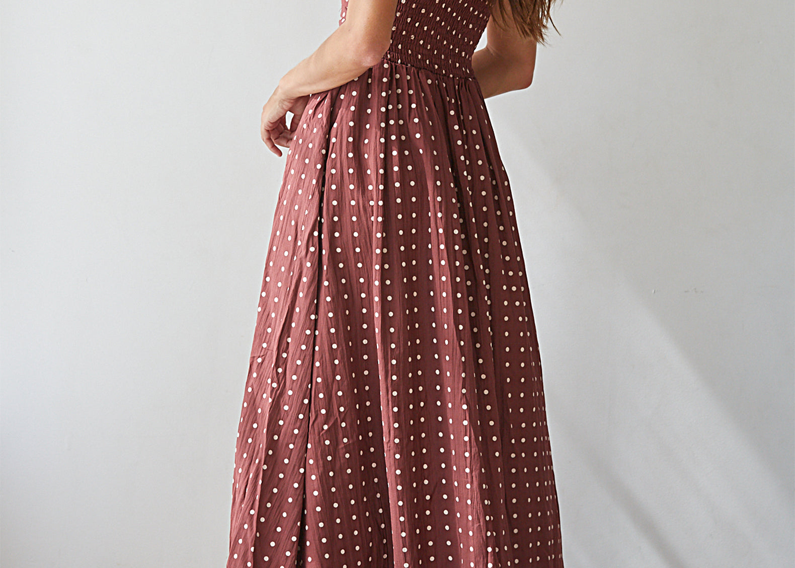 A woman wearing a strapless dusty rose maxi dress with white polka dots, shown from a rear three-quarter angle against a plain white wall.