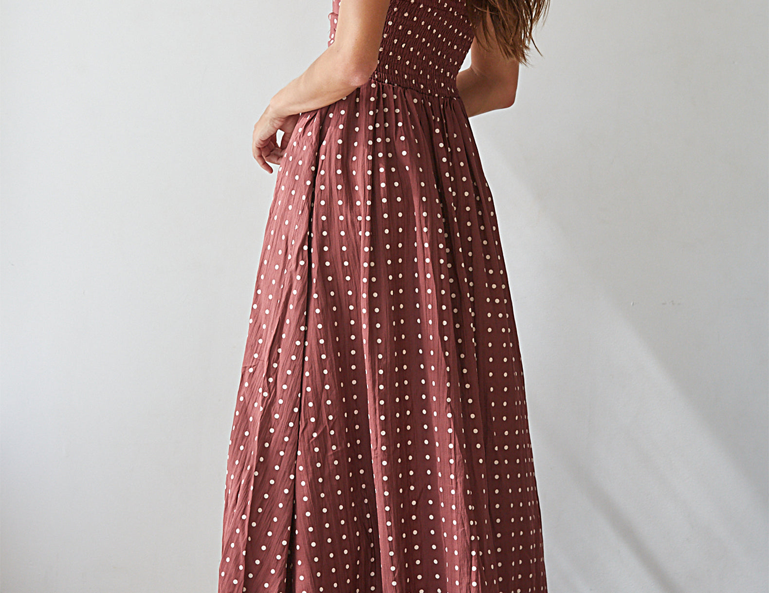 A woman wearing a strapless dusty rose maxi dress with white polka dots, shown from a rear three-quarter angle against a plain white wall.
