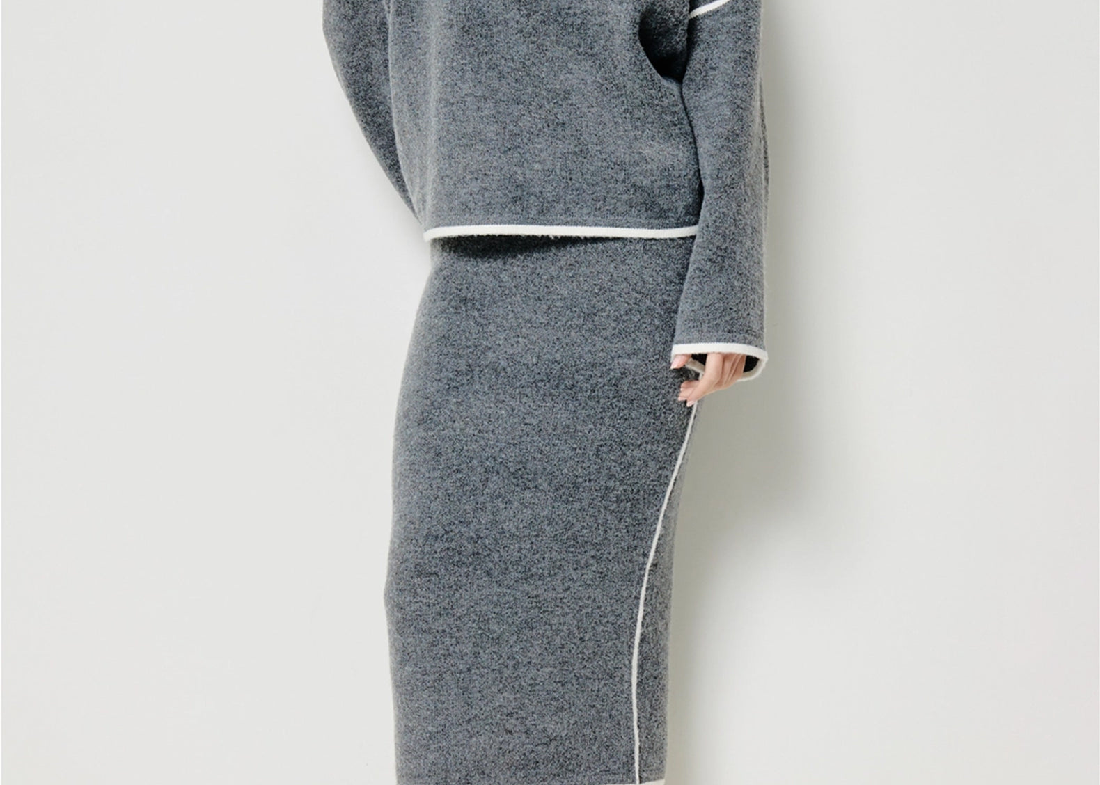 A model wearing a gray knit sweater and matching midi skirt set with white contrast trim, paired with off-white ankle boots, against a plain light gray studio background.