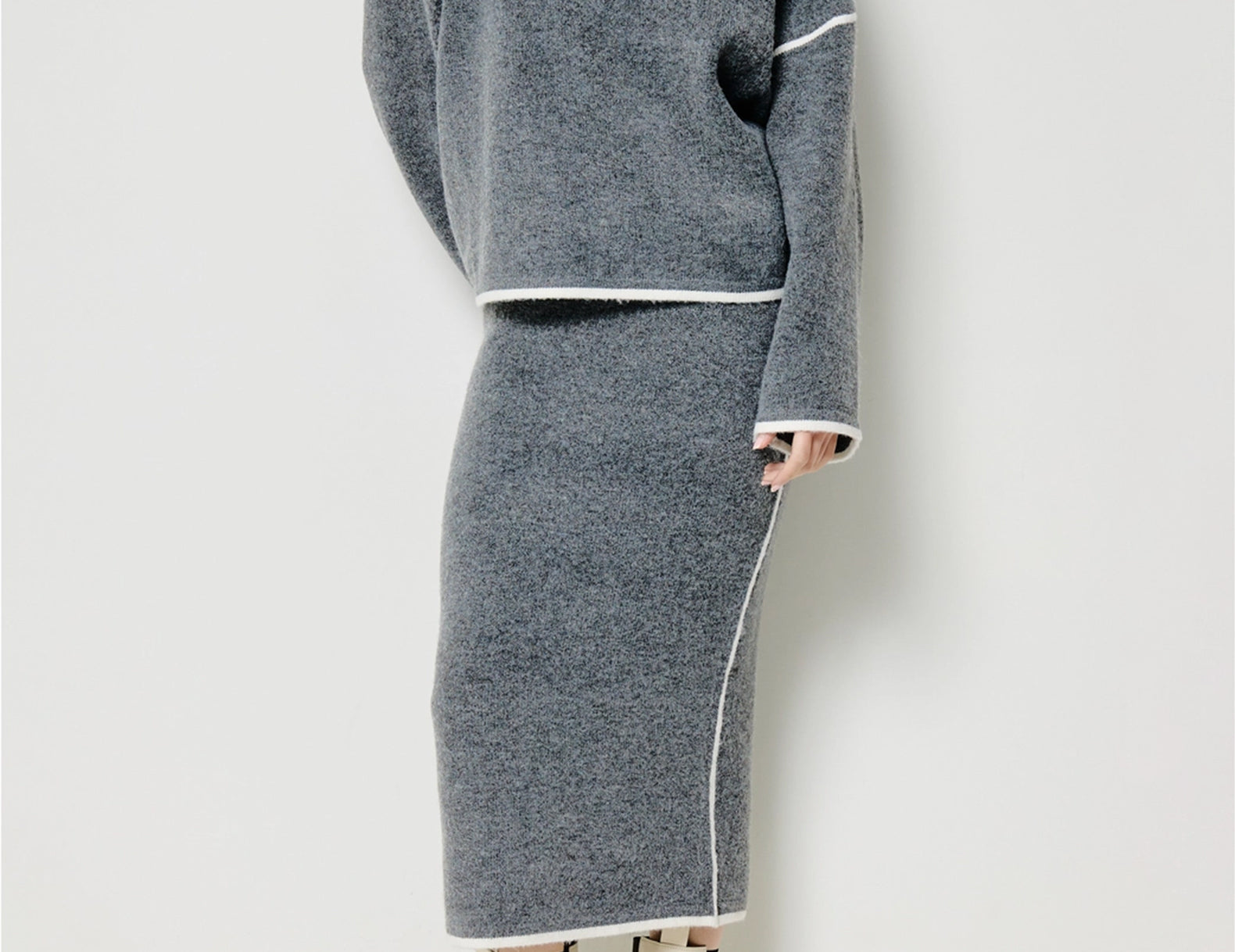 A model wearing a gray knit sweater and matching midi skirt set with white contrast trim, paired with off-white ankle boots, against a plain light gray studio background.