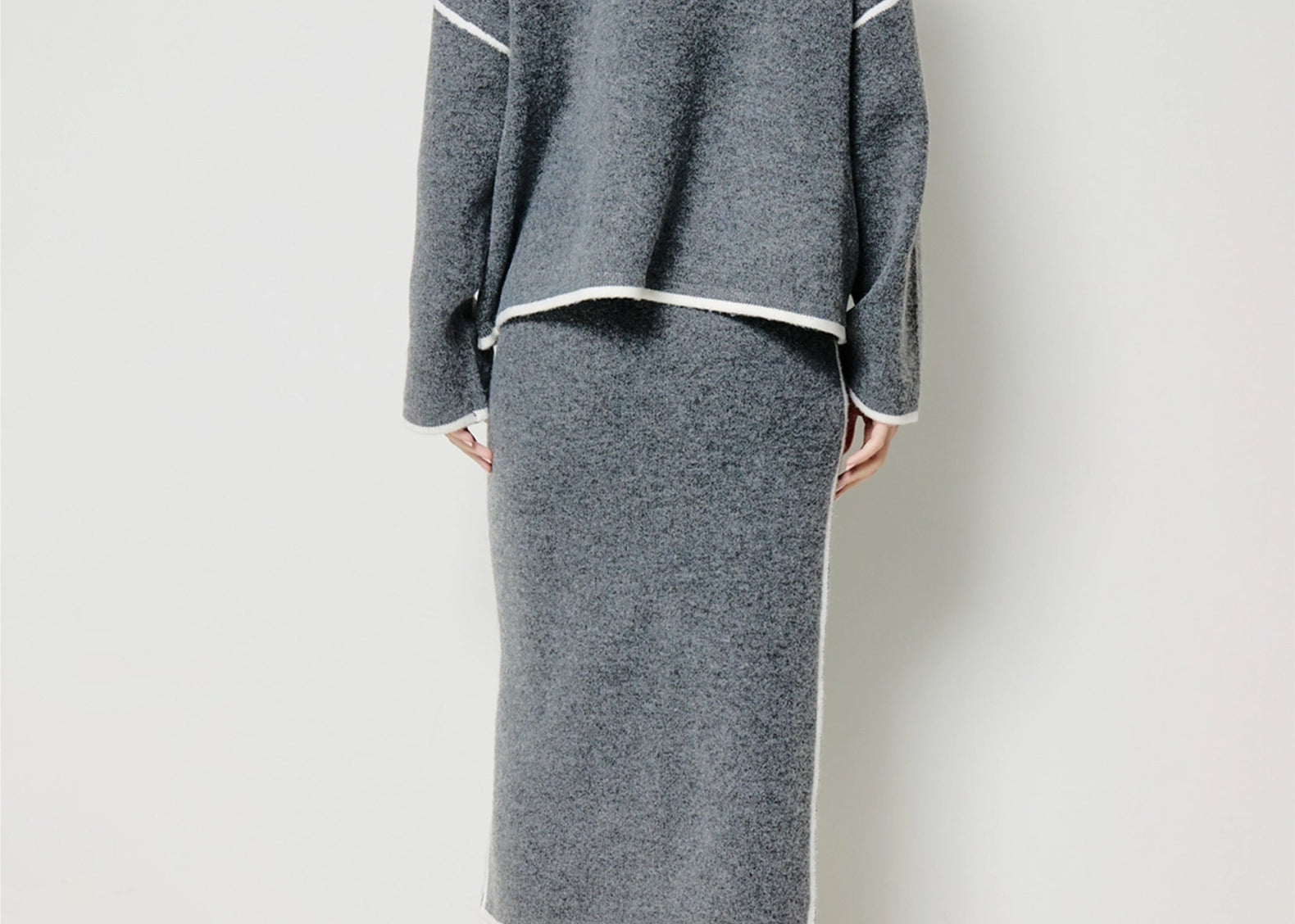 A model shown from the back wearing a gray knit two-piece set consisting of a cropped long-sleeve cardigan and a matching midi skirt, both with white contrast trim, paired with cream and black ankle boots against a plain white studio background.