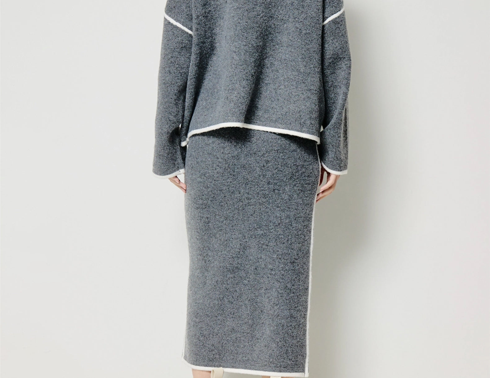 A model shown from the back wearing a gray knit two-piece set consisting of a cropped long-sleeve cardigan and a matching midi skirt, both with white contrast trim, paired with cream and black ankle boots against a plain white studio background.