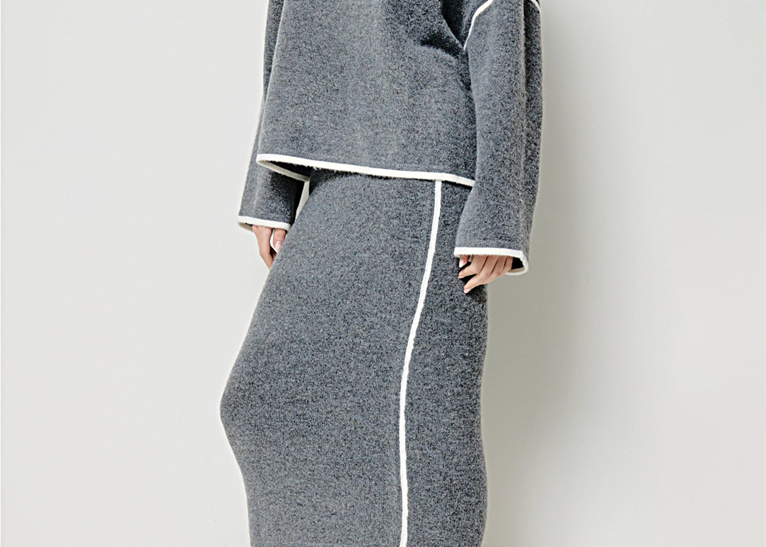 A model wearing a gray knit sweater and matching midi skirt set with white contrast piping, paired with cream Chelsea boots, against a light gray studio background.