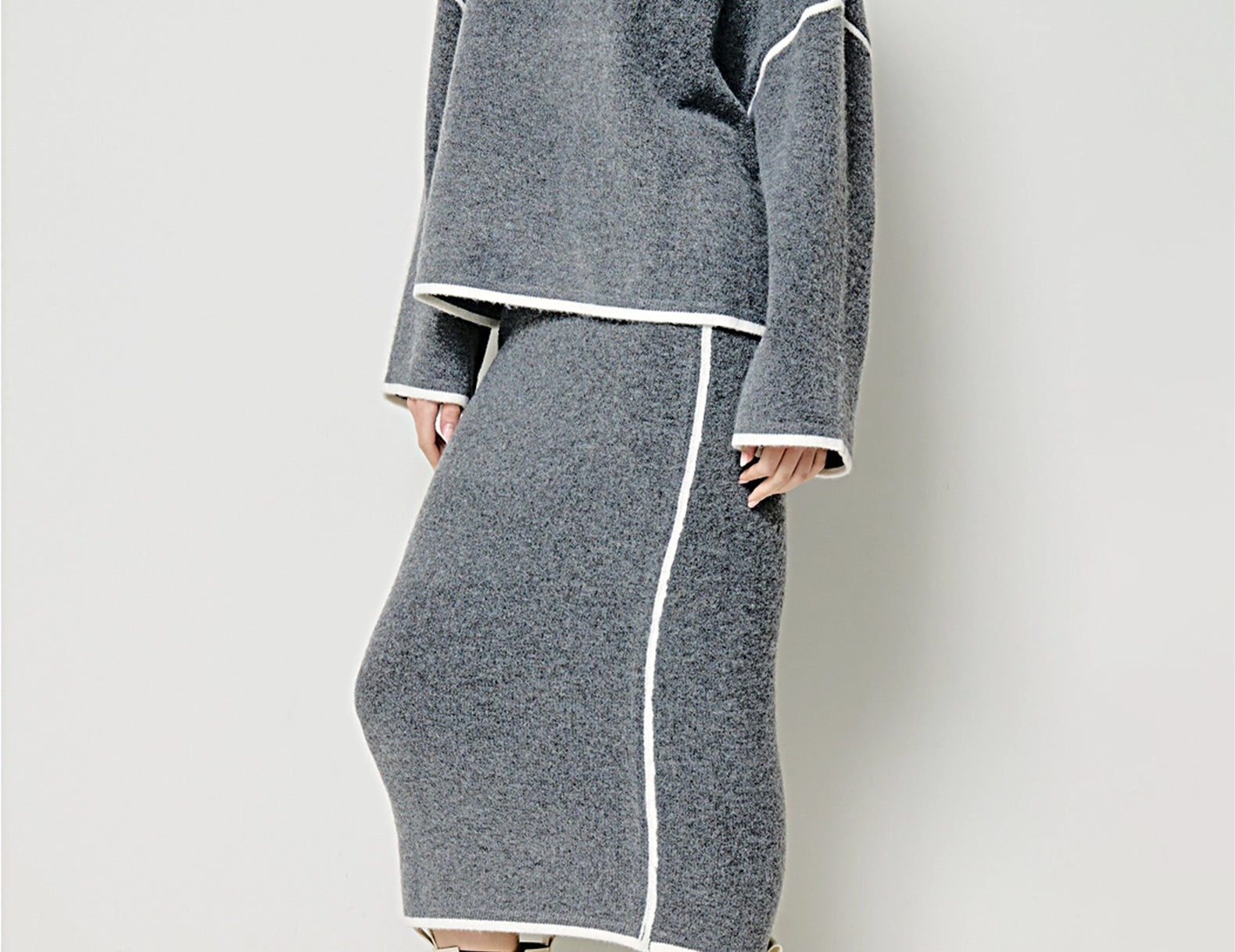A model wearing a gray knit sweater and matching midi skirt set with white contrast piping, paired with cream Chelsea boots, against a light gray studio background.