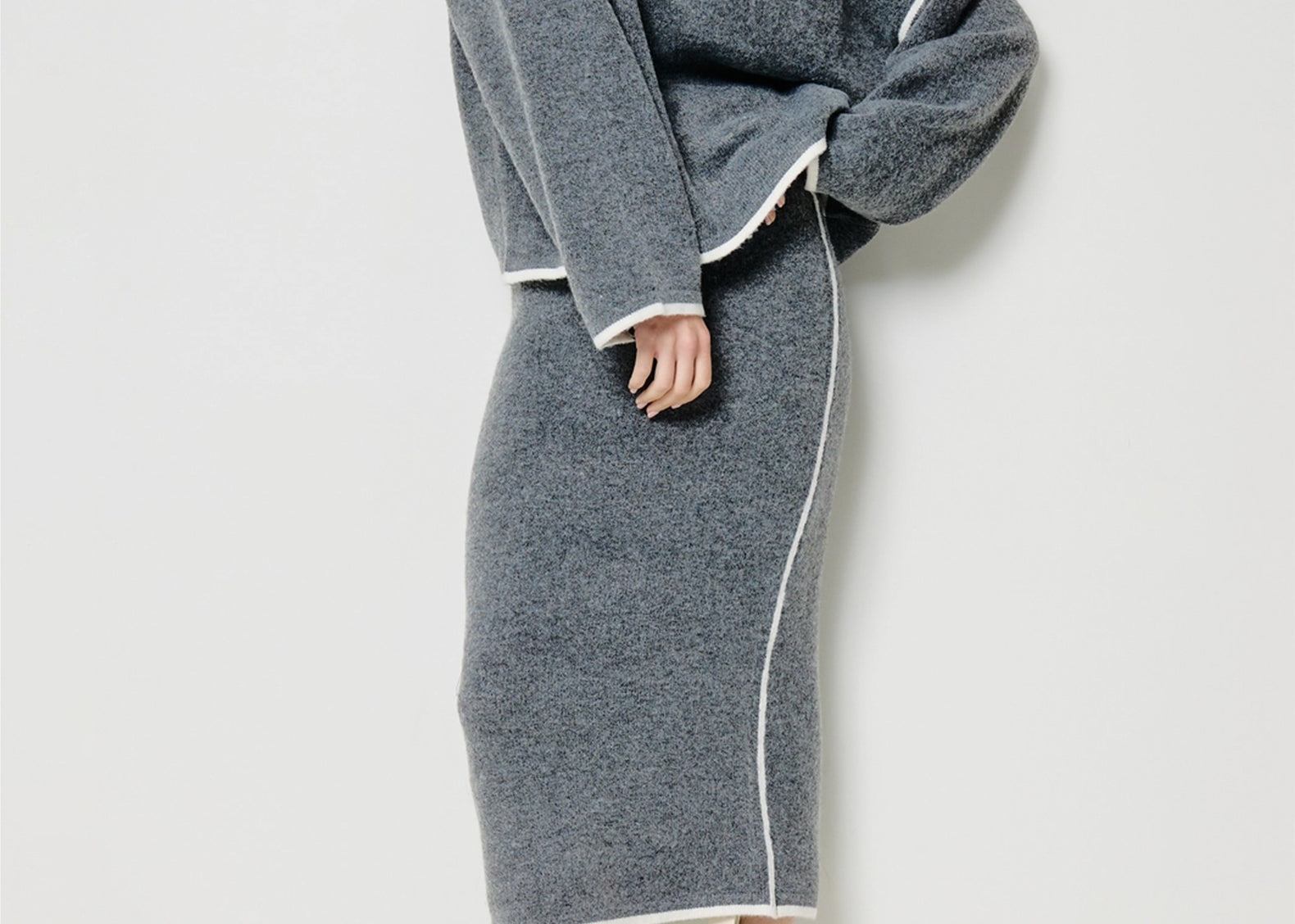 A model wears a matching gray knit sweater and midi skirt set with contrasting white piping, paired with cream ankle boots, against a plain light gray studio background.