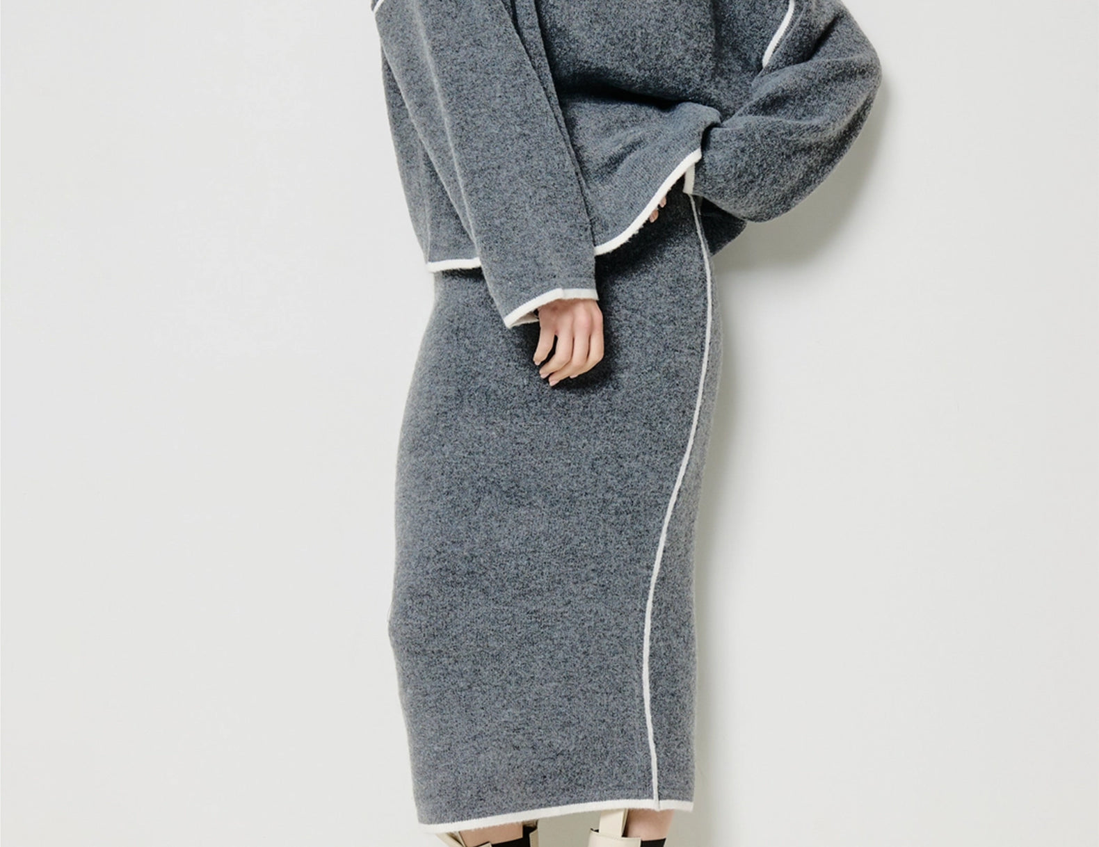 A model wears a matching gray knit sweater and midi skirt set with contrasting white piping, paired with cream ankle boots, against a plain light gray studio background.