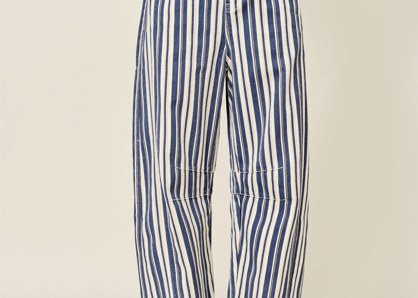 A person wearing loose, wide-leg striped pants with vertical blue and off-white stripes, paired with brown lace-up shoes, photographed against a plain light beige studio background.