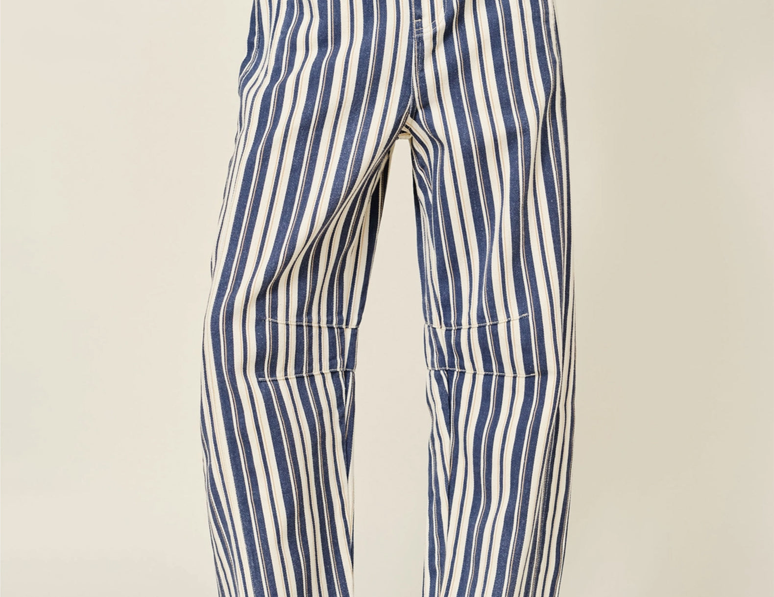 A person wearing loose vertical-striped barrel pants in blue and off-white, paired with brown leather loafers, photographed against a plain light beige studio background.