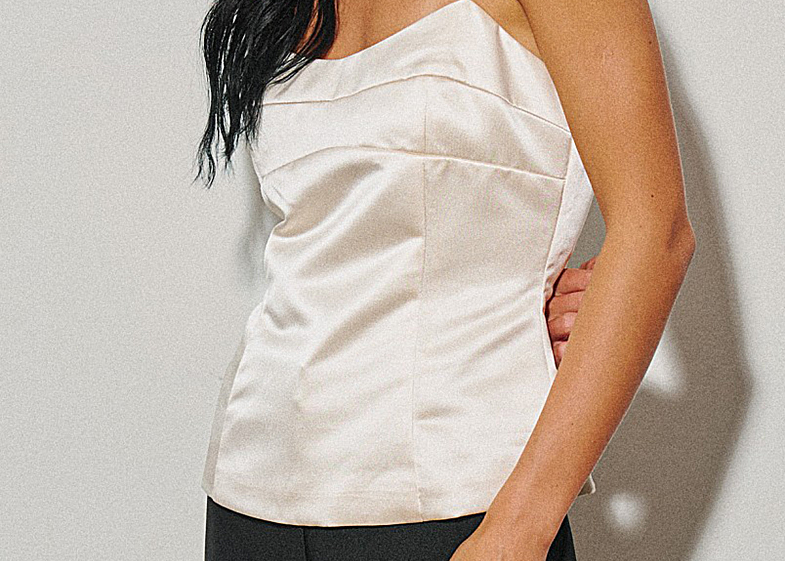 A woman wearing a cream satin camisole top with thin spaghetti straps and structured seams, paired with black trousers, set against a neutral light gray background.