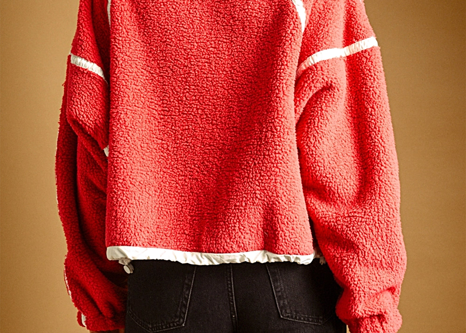 A person shown from the back wearing a textured red fleece jacket with white contrast stitching and black pants, set against a solid warm brown studio background.