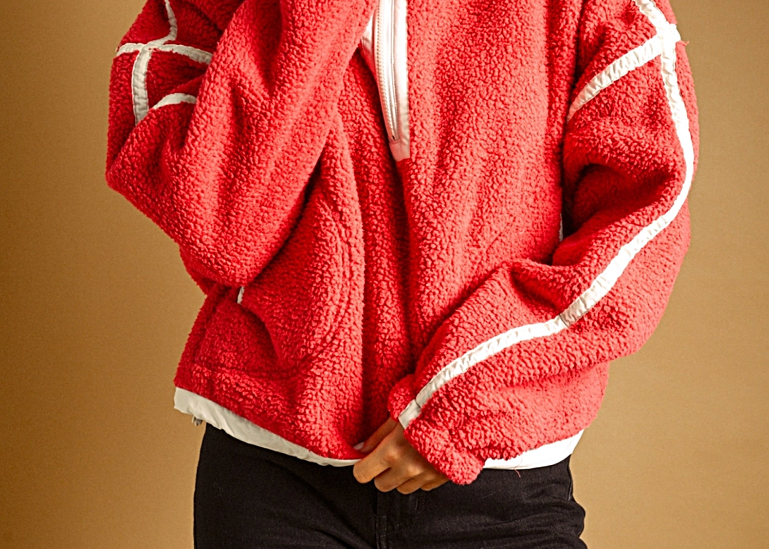 A woman wearing a bright red sherpa fleece zip-up jacket with white contrast stripes and black pants, posed against a solid warm brown background.