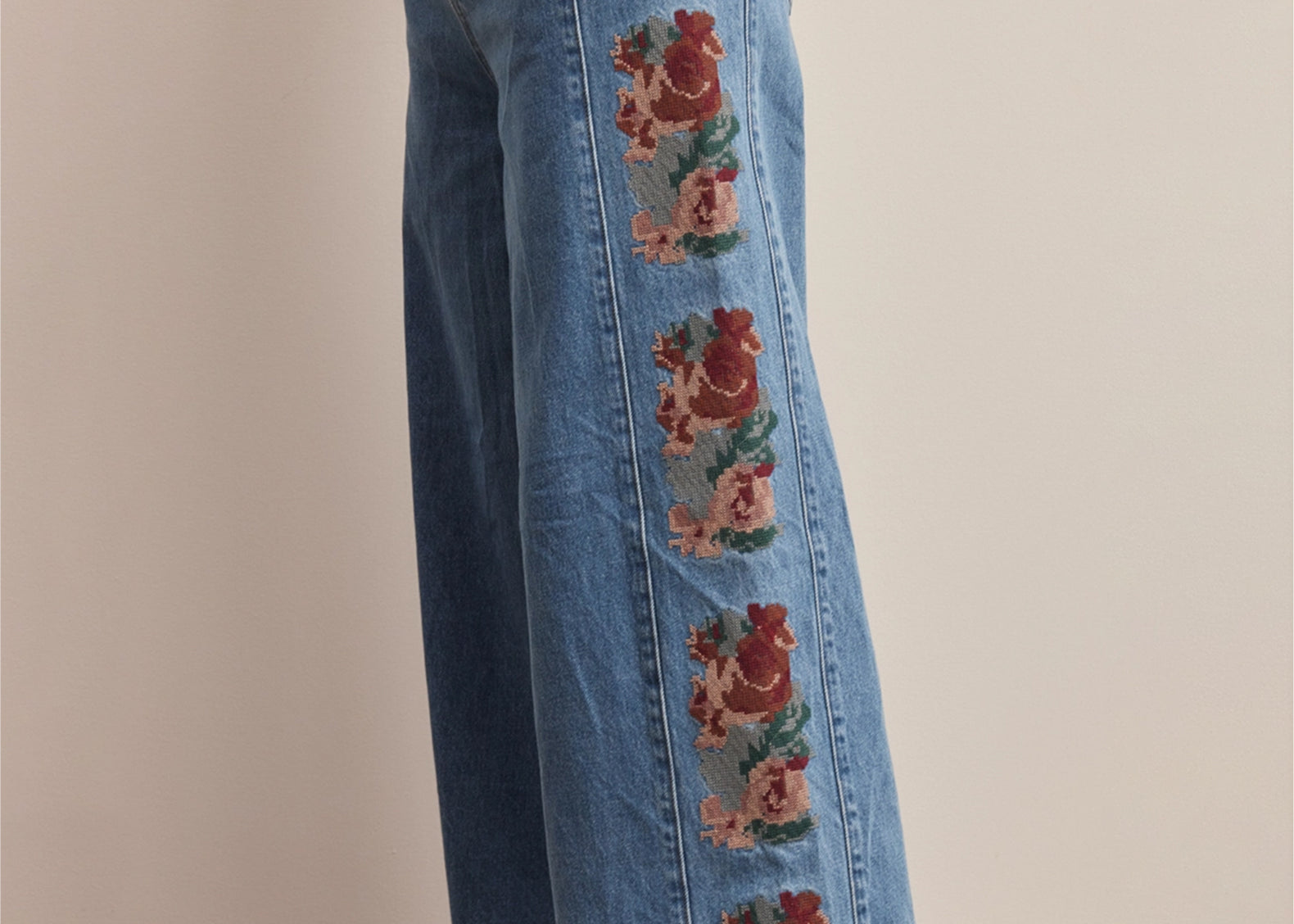 A person wearing wide-leg blue denim jeans with vertical floral embroidery down the outer side, paired with beige flats, standing against a plain light beige wall.