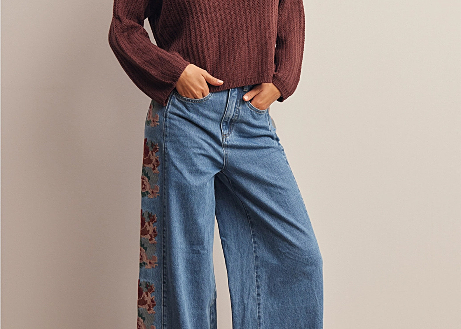 A woman models a dark brown ribbed knit sweater and wide-leg blue jeans with a floral patterned side panel, standing against a neutral beige wall.