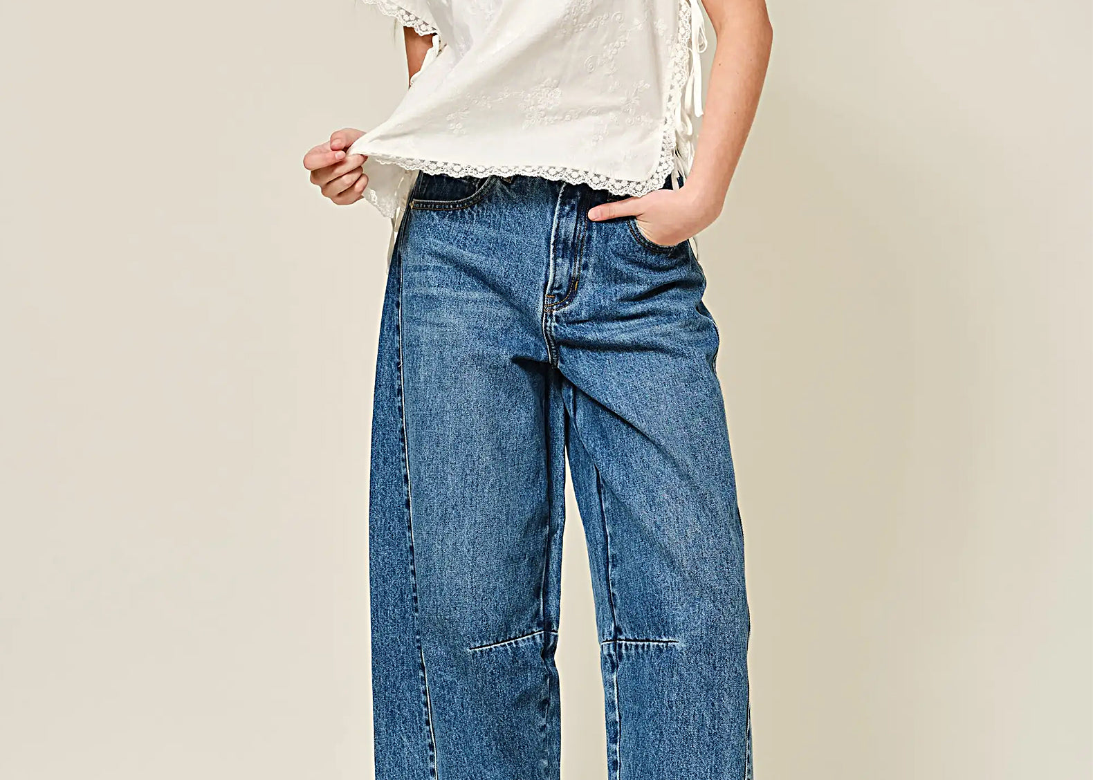 A model wearing a white short-sleeve top with lace trim, blue cropped wide-leg denim jeans, and brown leather loafers, standing against a solid light beige studio background.