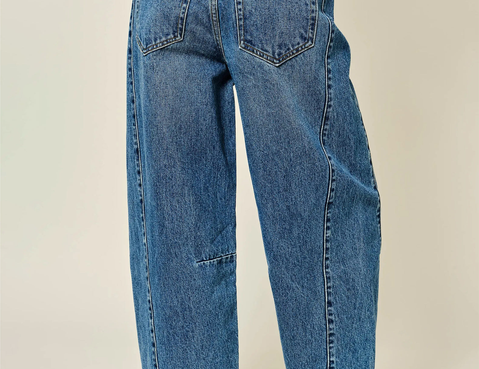 Rear view of a person wearing wide-leg blue denim jeans and brown leather loafers against a plain light beige studio background.