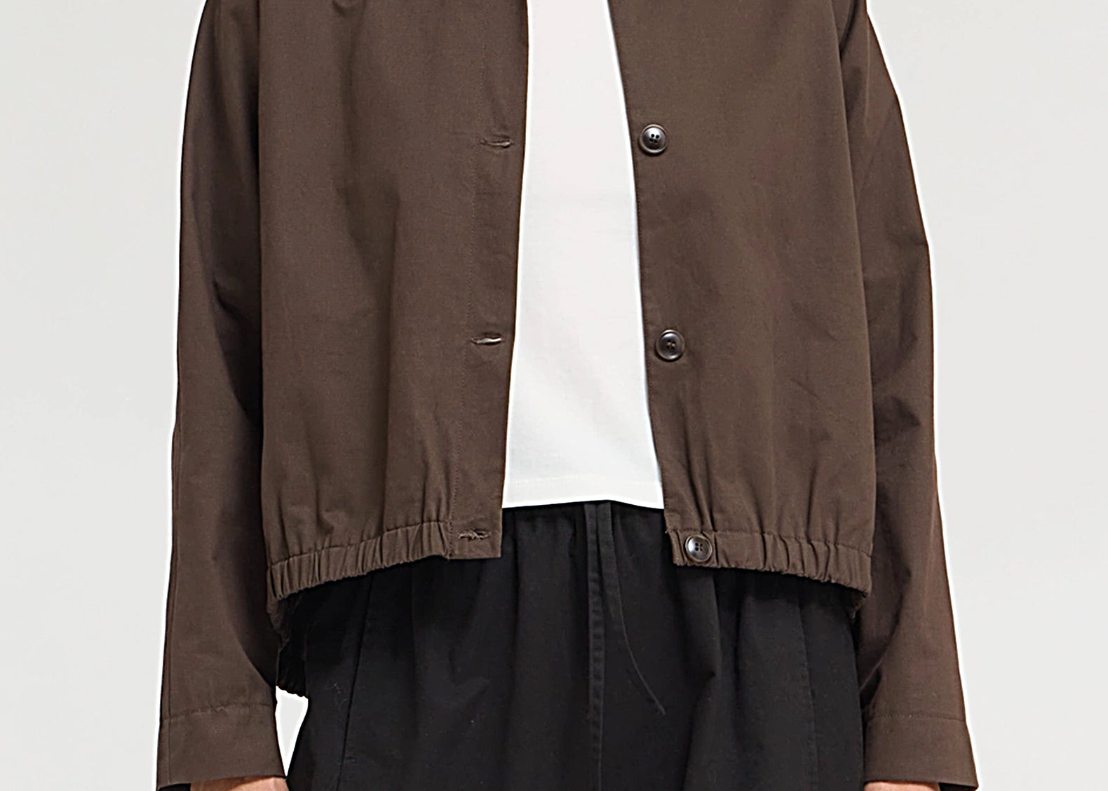 A model wearing a dark brown cropped jacket with a funnel collar and snap-button front, layered over a white top and black wide-leg pants, against a light gray studio background.