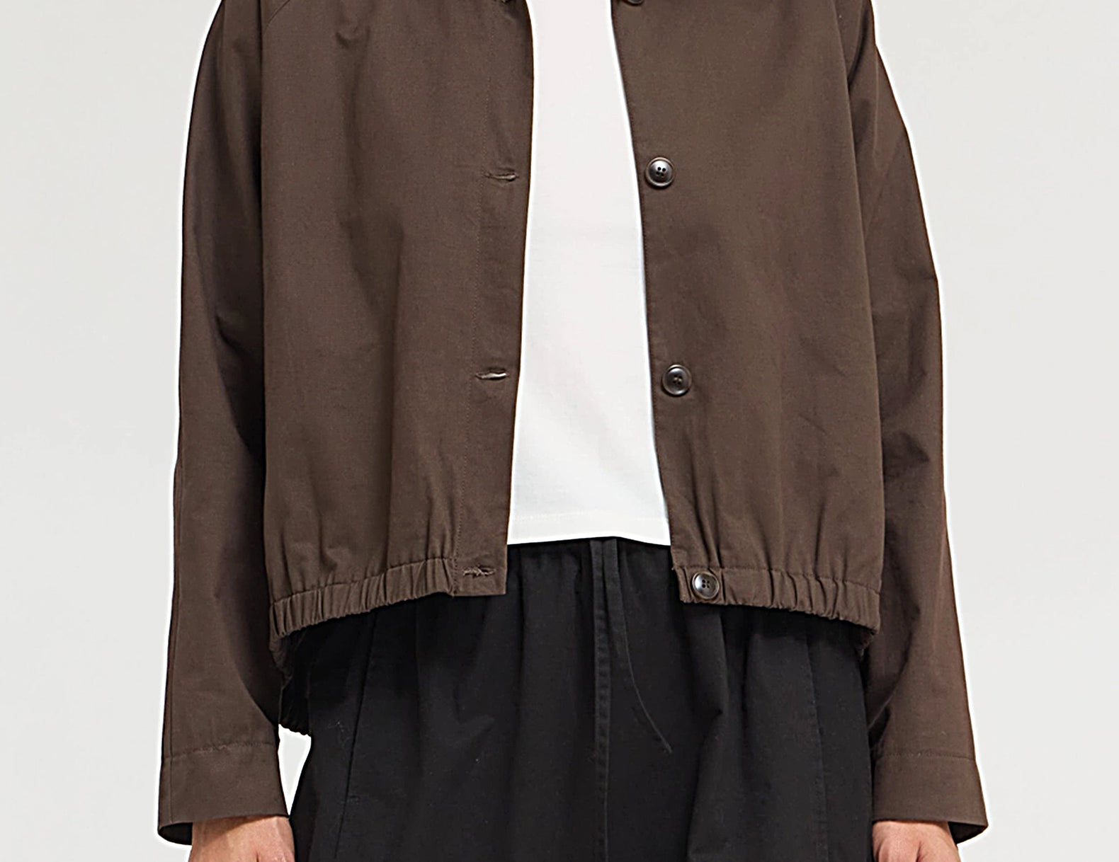 A model wearing a dark brown cropped jacket with a funnel collar and snap-button front, layered over a white top and black wide-leg pants, against a light gray studio background.