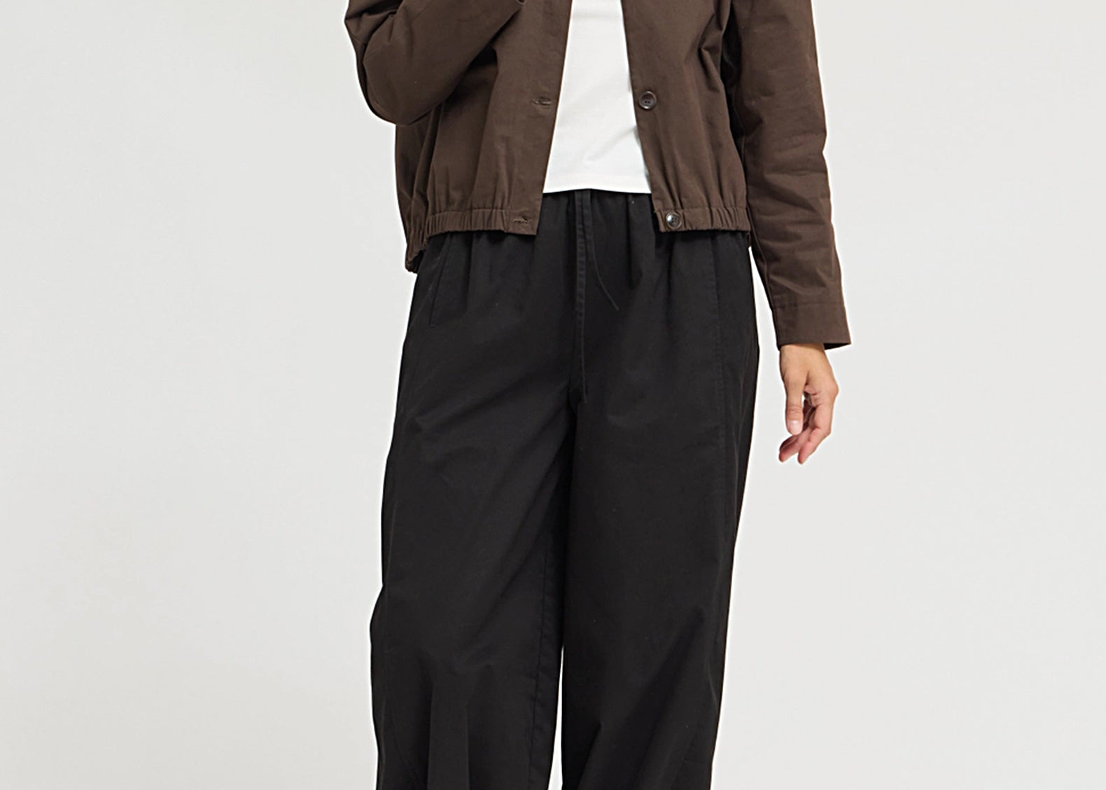 A model wearing a brown cropped button-up jacket over a white t-shirt, paired with loose black wide-leg pants and black shoes, photographed against a light gray studio background.