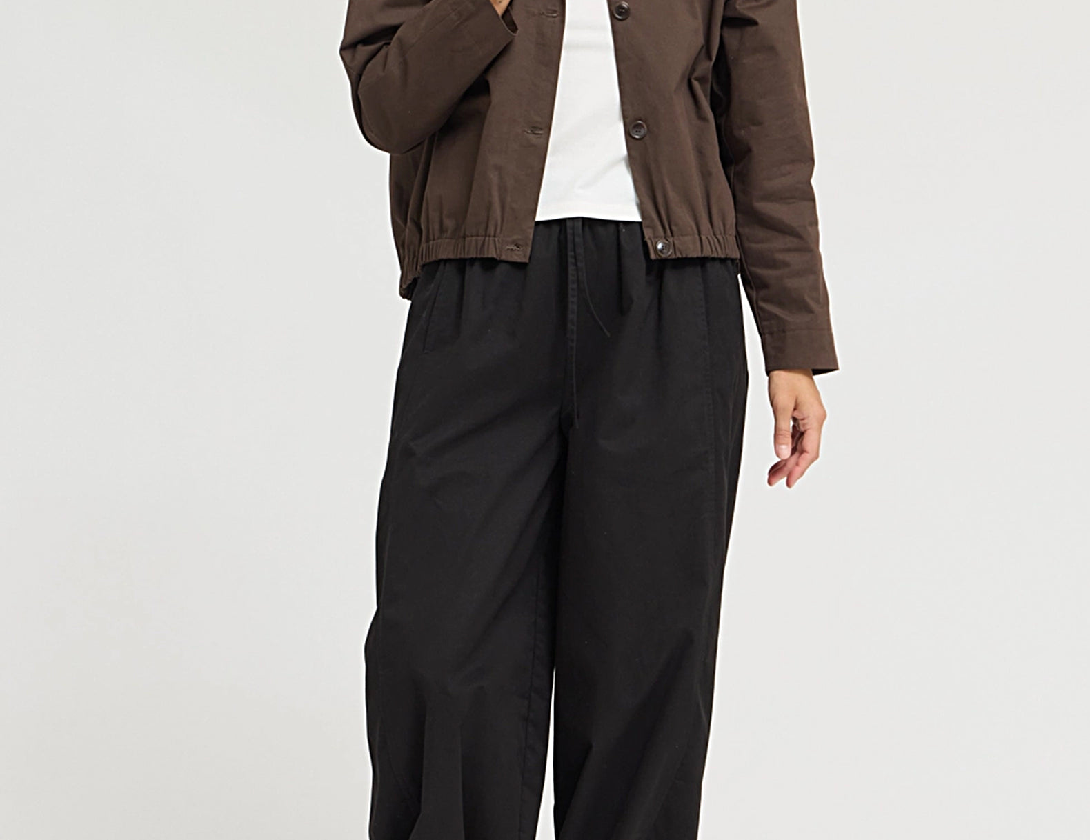 A model wearing a brown cropped button-up jacket over a white t-shirt, paired with loose black wide-leg pants and black shoes, photographed against a light gray studio background.