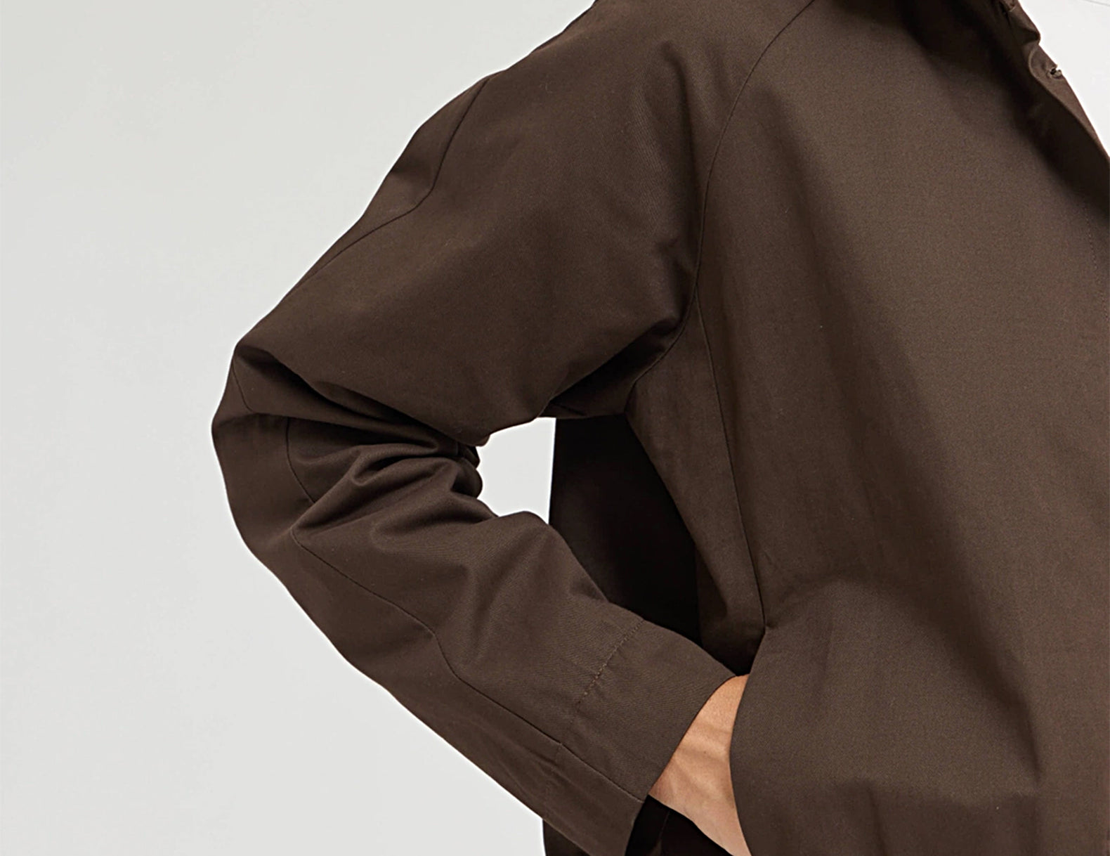 A close-up of a person wearing a dark brown lightweight jacket with a high collar, elastic hem, and long sleeves, paired with a white shirt underneath.