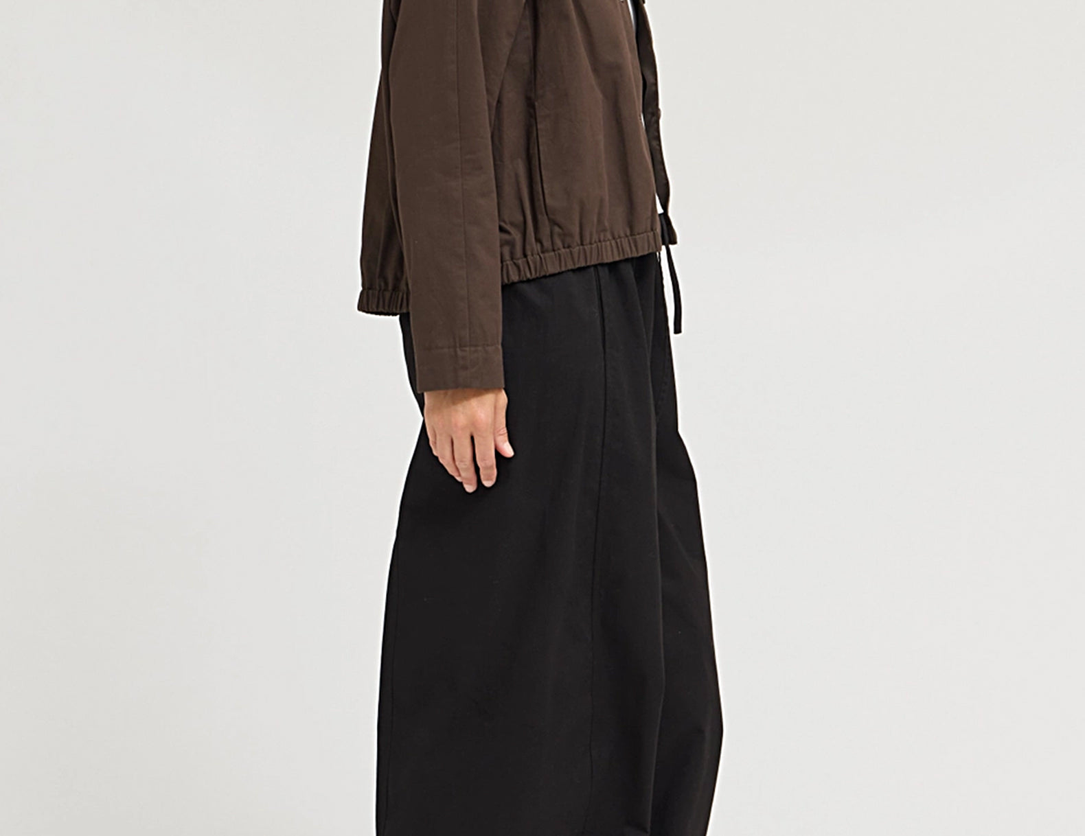 A model shown from the side wearing a dark brown cropped utility jacket and loose black wide-leg pants against a plain light gray studio background.