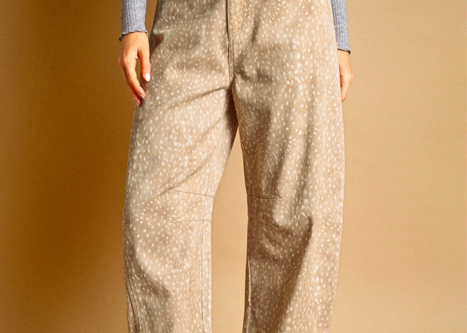 A person wearing loose beige pants with a small white speckled fawn pattern, paired with a gray ribbed long-sleeve top and dark brown sneakers, standing against a warm tan studio background.