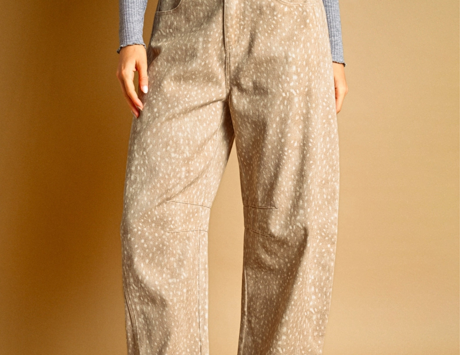 A person wearing loose beige pants with a small white speckled fawn pattern, paired with a gray ribbed long-sleeve top and dark brown sneakers, standing against a warm tan studio background.