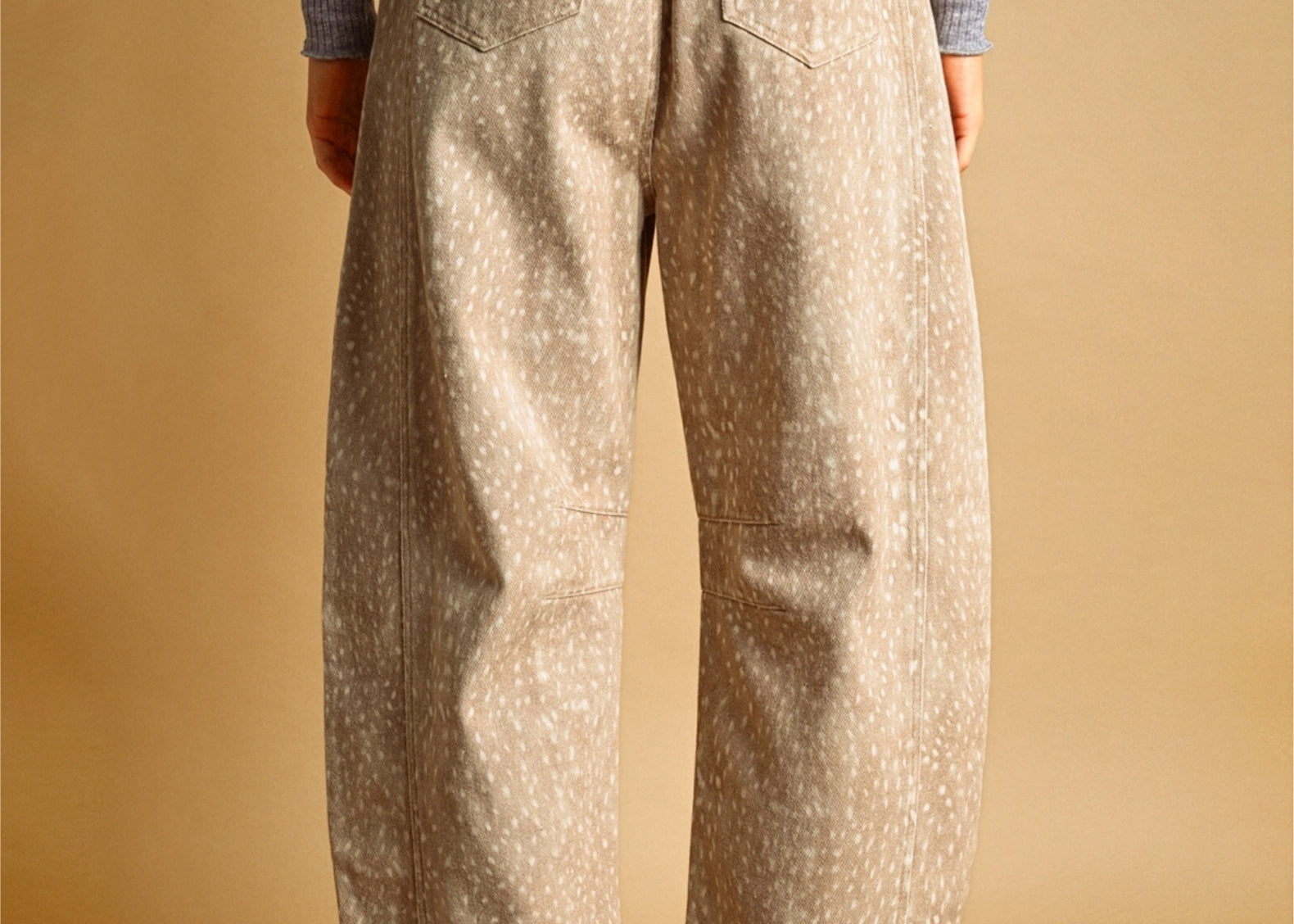 A person shown from the back wearing loose, wide-leg beige pants with a small white speckled fawn pattern, a gray ribbed long-sleeve top, and brown-and-white sneakers against a warm tan studio background.