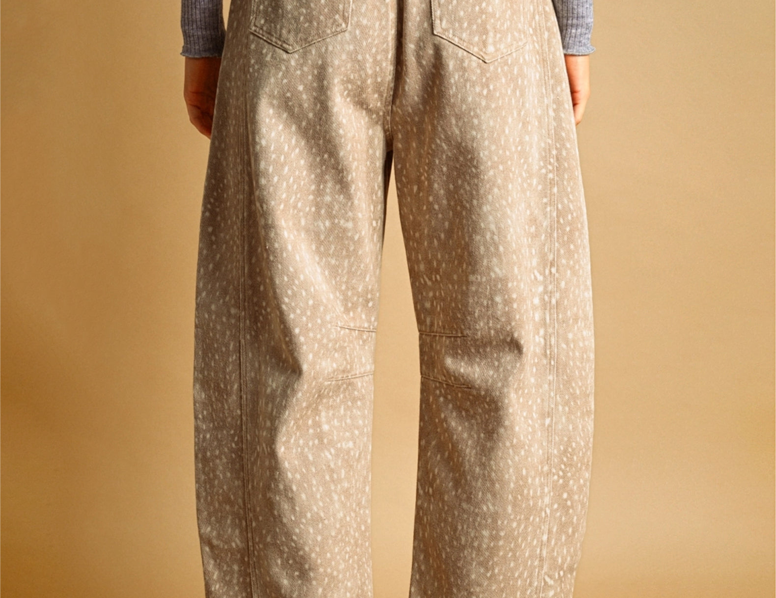 A person shown from the back wearing loose, wide-leg beige pants with a small white speckled fawn pattern, a gray ribbed long-sleeve top, and brown-and-white sneakers against a warm tan studio background.