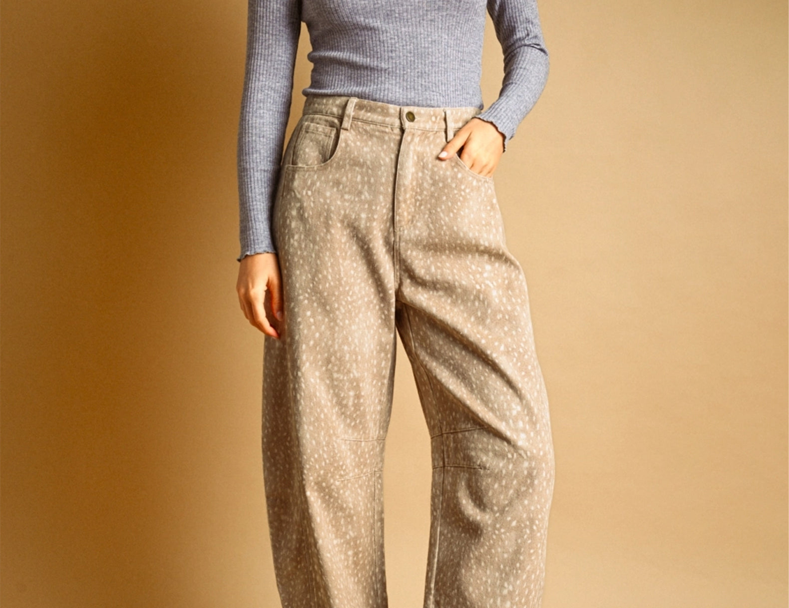 A model wearing a gray long-sleeve knit top and beige wide-leg fawn-patterned trousers, paired with brown sneakers, standing against a warm beige studio backdrop.