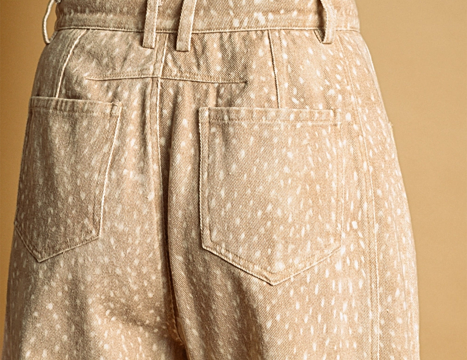 Close-up back view of a person wearing beige wide-leg trousers with a white speckled fawn pattern, paired with a gray ribbed sweater, against a warm beige background.