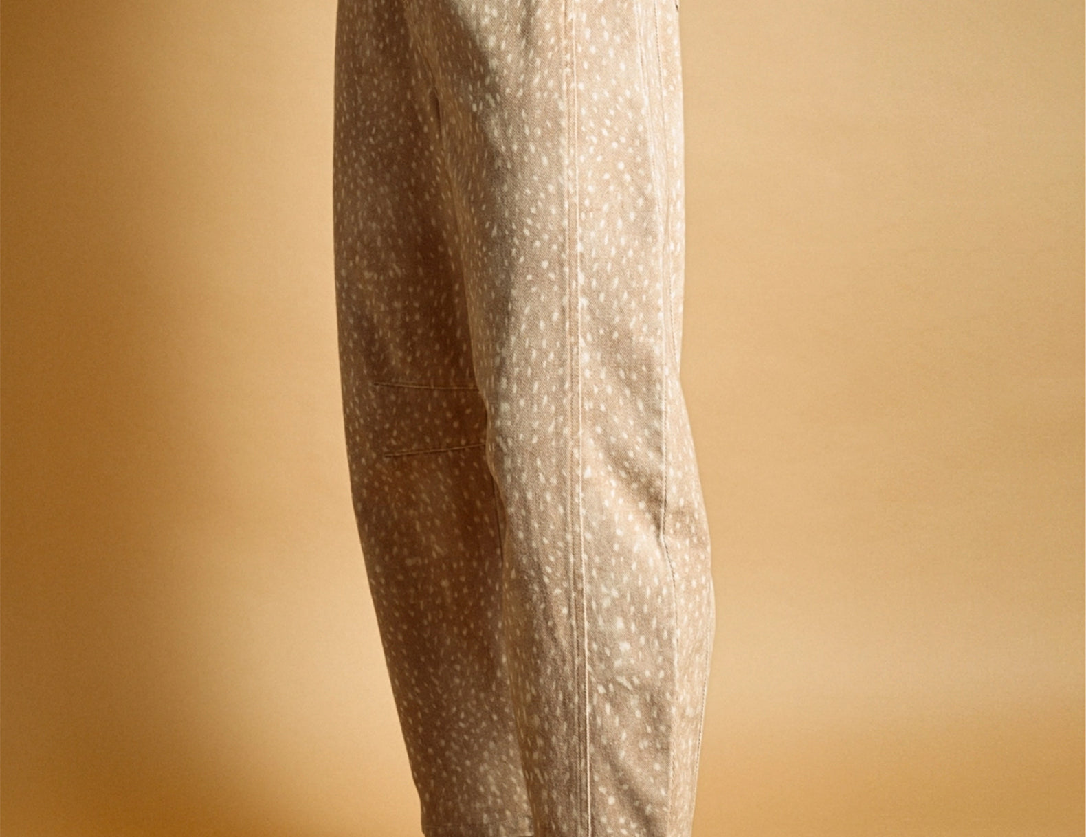 A person shown from the waist down wearing loose, wide-leg beige trousers with a small white speckled fawn pattern, paired with brown suede sneakers and white socks, against a warm tan studio background.