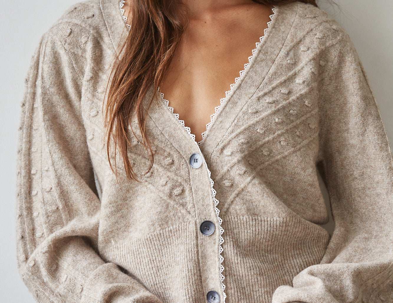 A woman wearing a beige knit cardigan with textured bobble details, scalloped white trim along the V-neck and button placket, and blue jeans, photographed against a plain light gray wall.