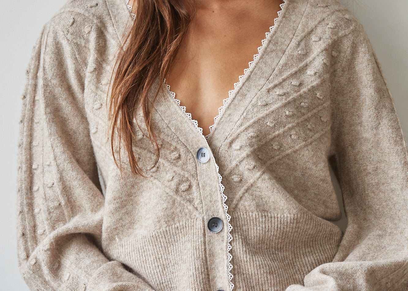 A woman wearing a beige knit cardigan with textured bobble details, scalloped white trim along the V-neck and button placket, and blue jeans, photographed against a plain light gray wall.