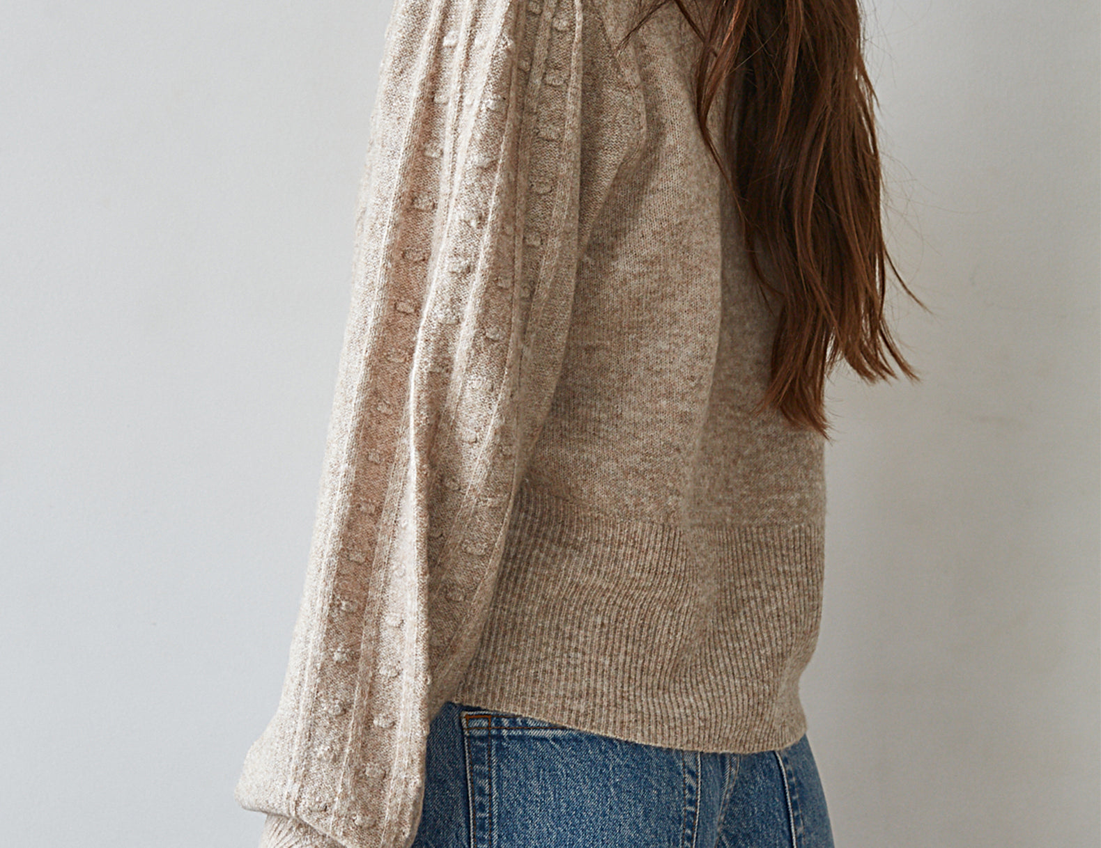 A woman wearing a beige textured knit sweater with dramatic balloon sleeves, shown from a side-back angle against a plain light gray background.