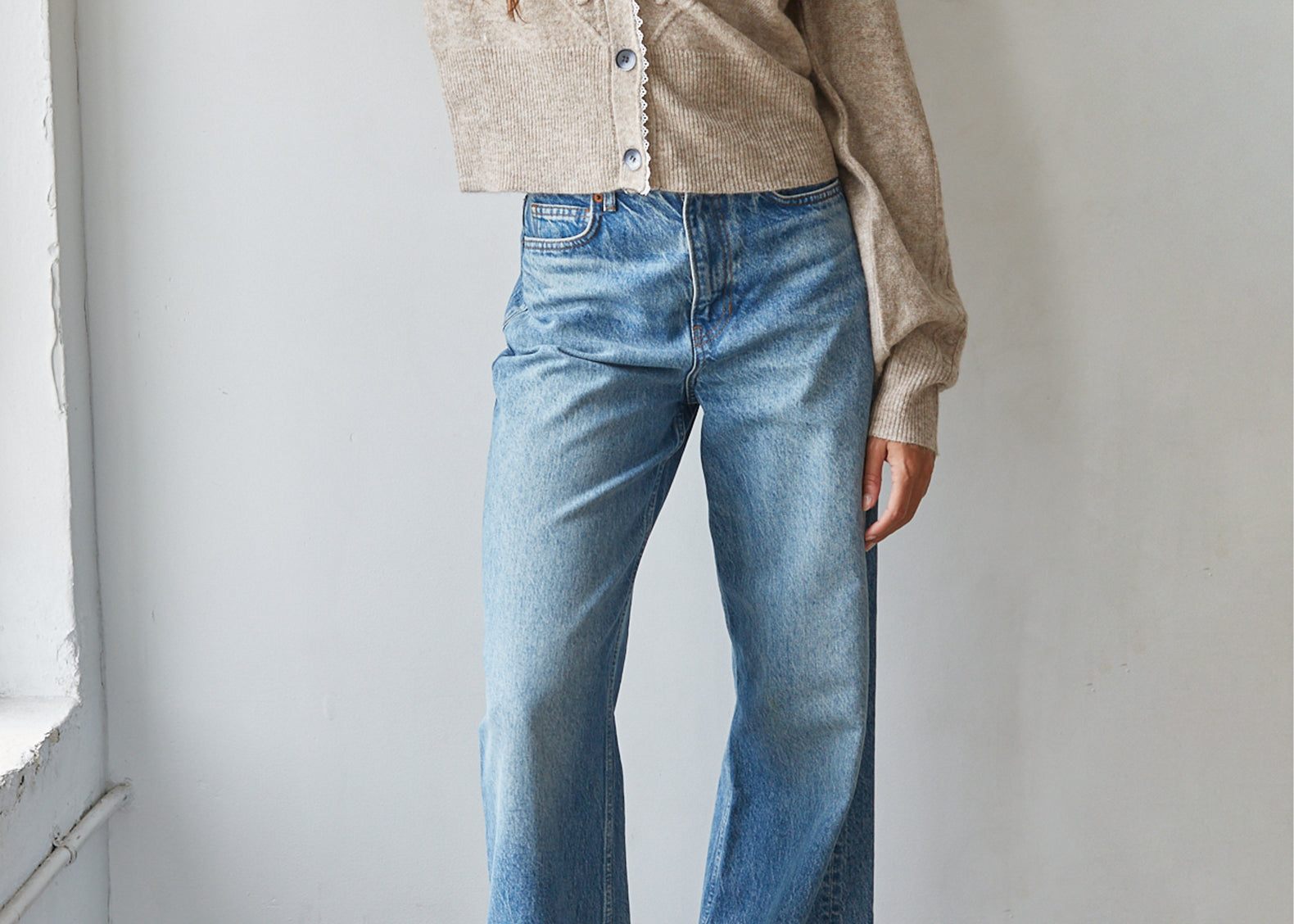 A woman wearing a beige knit cropped cardigan and light blue wide-leg jeans, standing against a white wall.