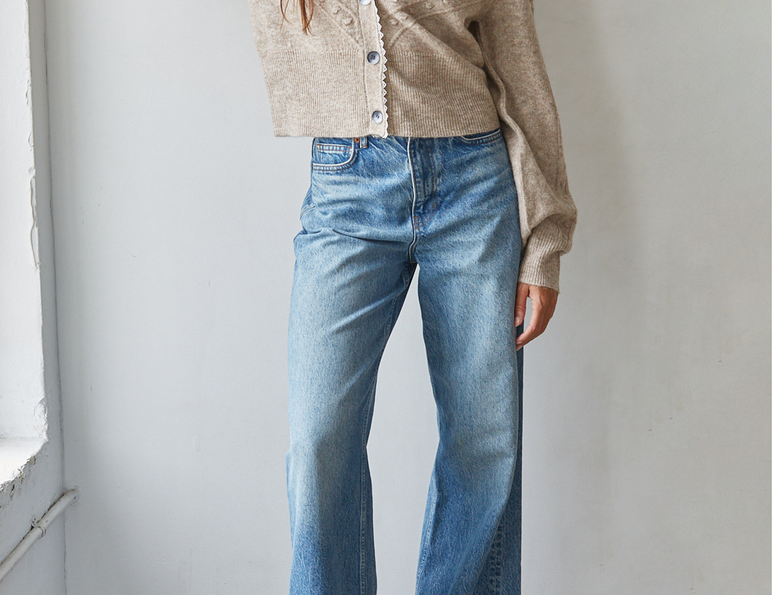 A woman wearing a beige knit cropped cardigan and light blue wide-leg jeans, standing against a white wall.