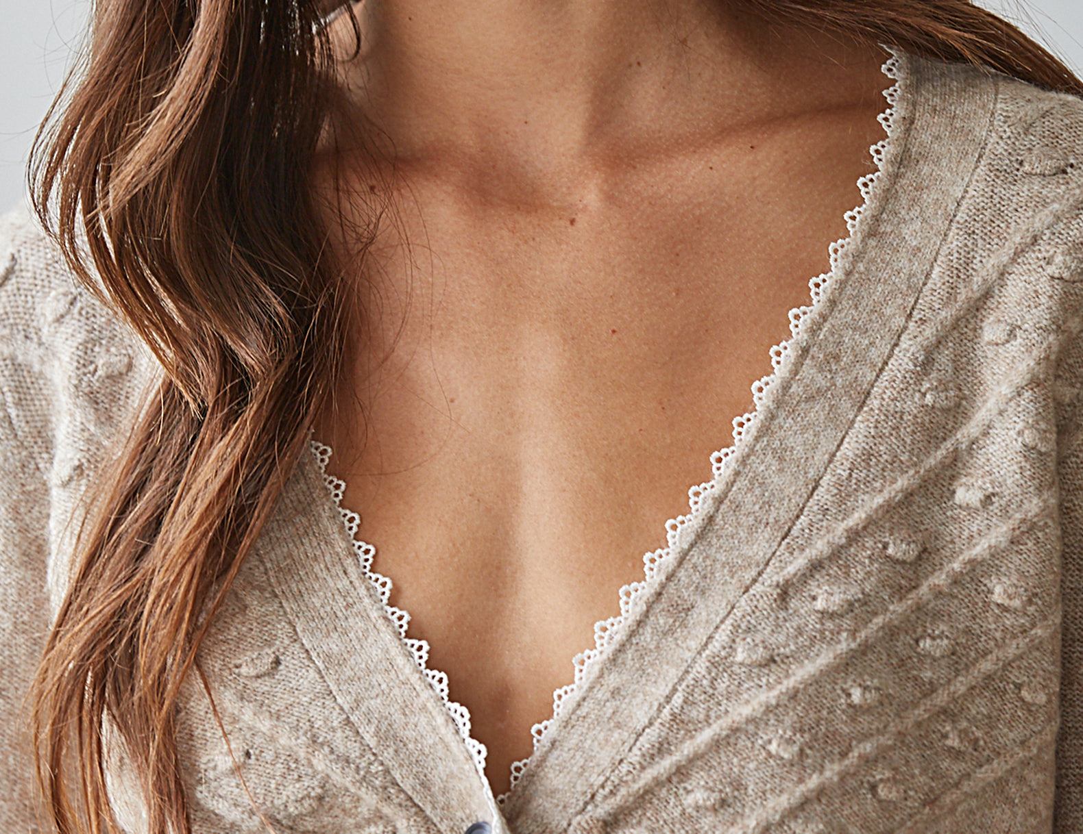Close-up of a woman wearing a light beige textured knit cardigan with a scalloped white lace trim along the V-neckline.