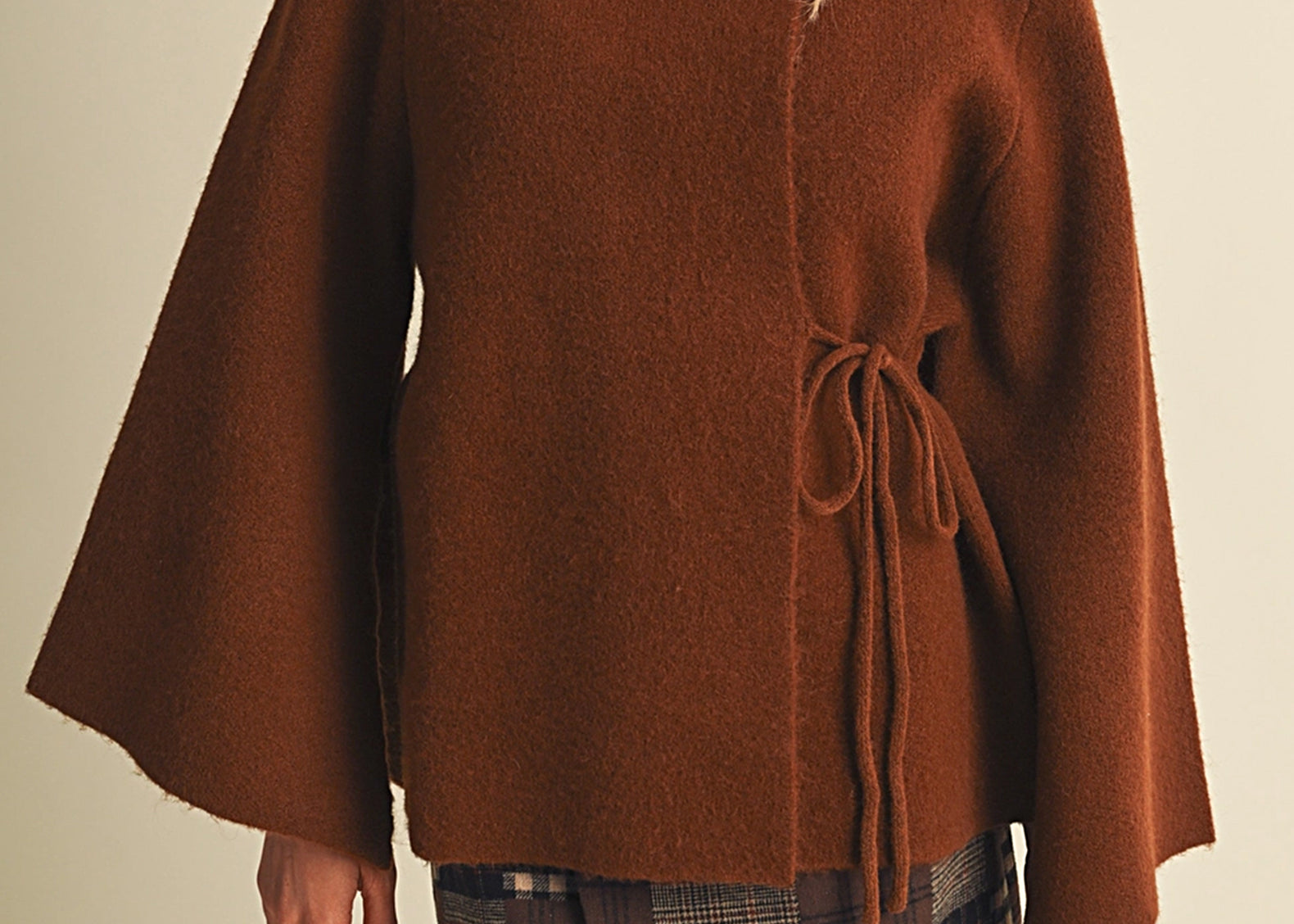 A woman wearing a brown fuzzy cape-style sweater with a drawstring waist and a plaid mini skirt, photographed against a plain light beige studio background.