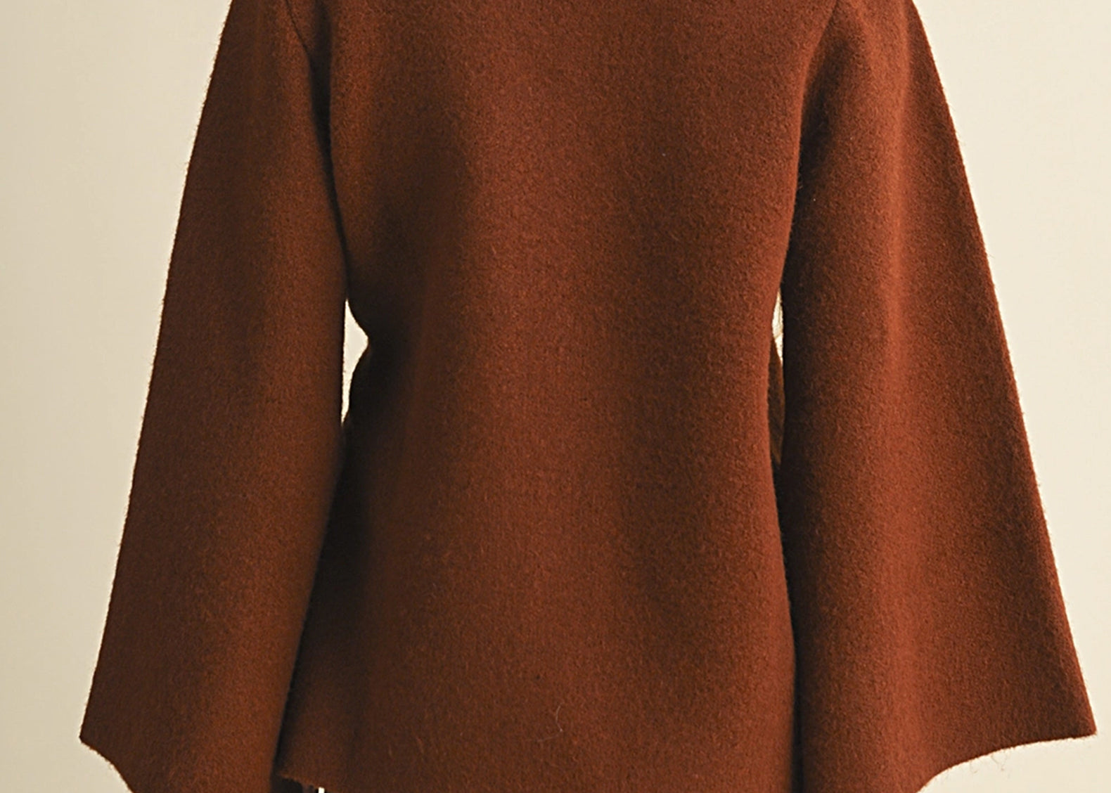 A woman shown from the back wearing a rust-brown long-sleeve cropped coat or cape with a fuzzy texture, paired with a plaid mini skirt, against a plain beige background.