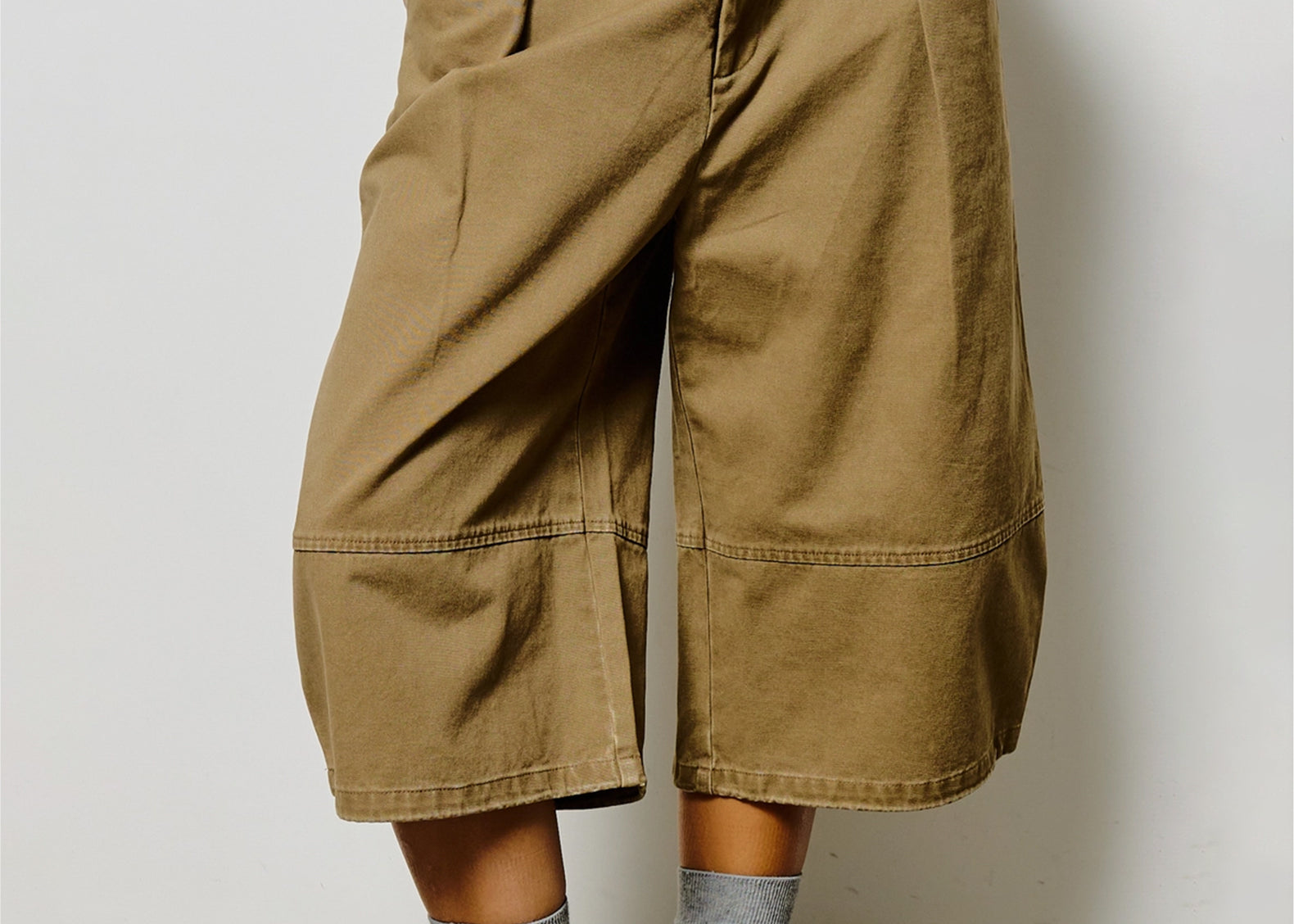 A person wearing oversized tan cropped pants with side pockets, paired with gray socks and patterned sneakers, standing against a plain light wall.